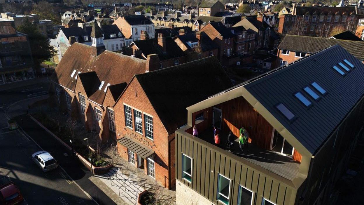 A drone photograph of the building shows the new, modern extension next to the old red brick building, which looks like a church, or an old school. The new building features a dark green metal roof and a balcony.