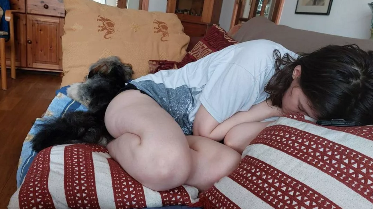 A woman with fair skin and brown hair laying asleep on a sofa, with her head buried in a red and white pillow. A fluffy dark grey and white dog is beside her.
