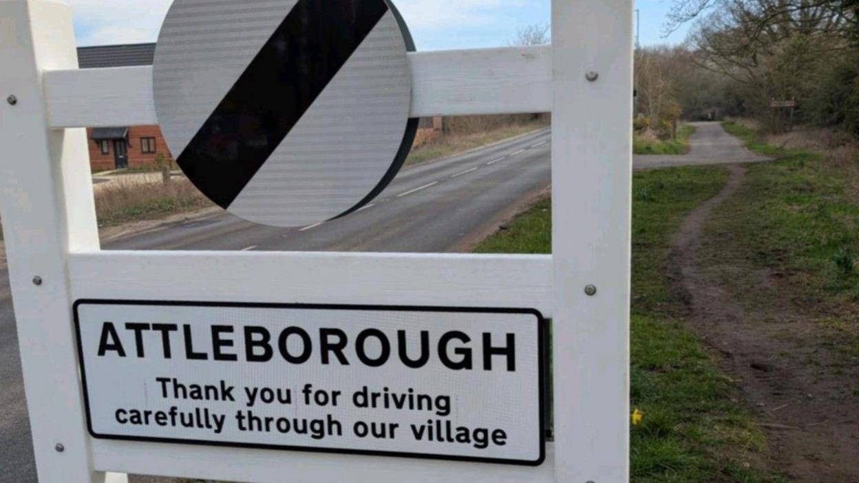 Road sign in black and white with a national speed limit marking and words saying "Attleborough Thank you for driving carefully through our village."