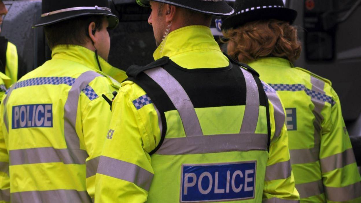 Three police officers in yellow high-visibility coats, with their backs to the camera