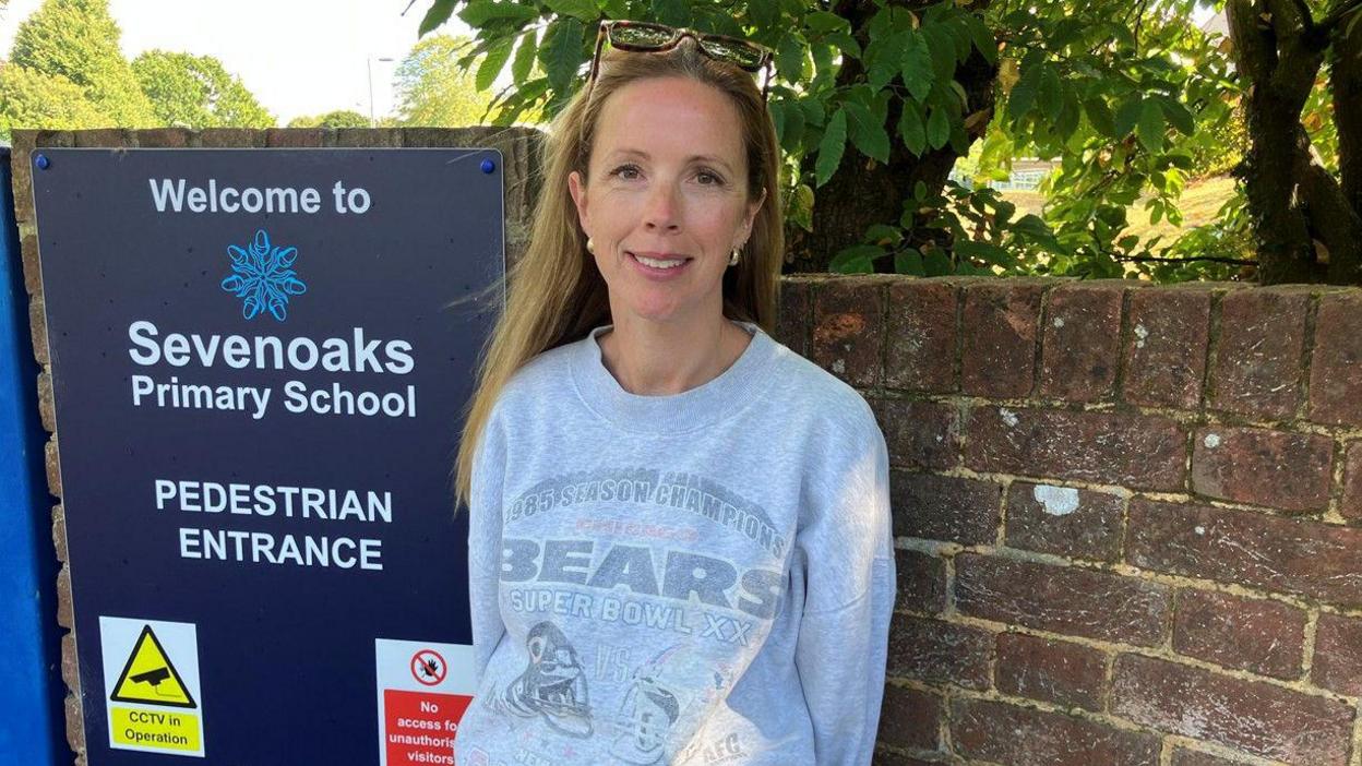 A woman in her early forties with long blonde hair smiles a the camera whilst wearing a grey sweatshirt. She stands next to a dark blue sign which reads : "Sevenoaks Primary School".