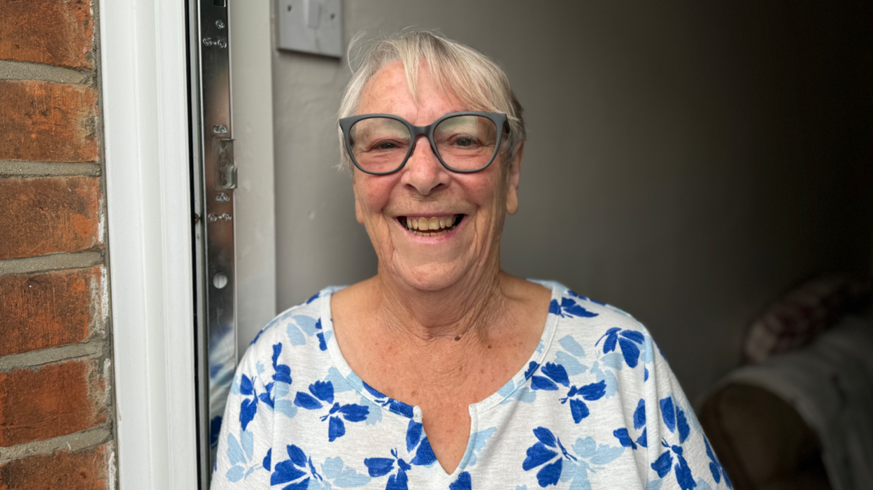 Jean McSorley wearing a blue and white floral top and glasses smiling into the camera while standing in her doorway.