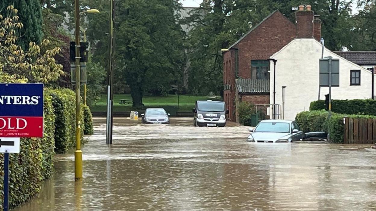 A flooded street in Horncastle showing a car partially submerged in water. A car and a van can be seen in the background next to two houses.