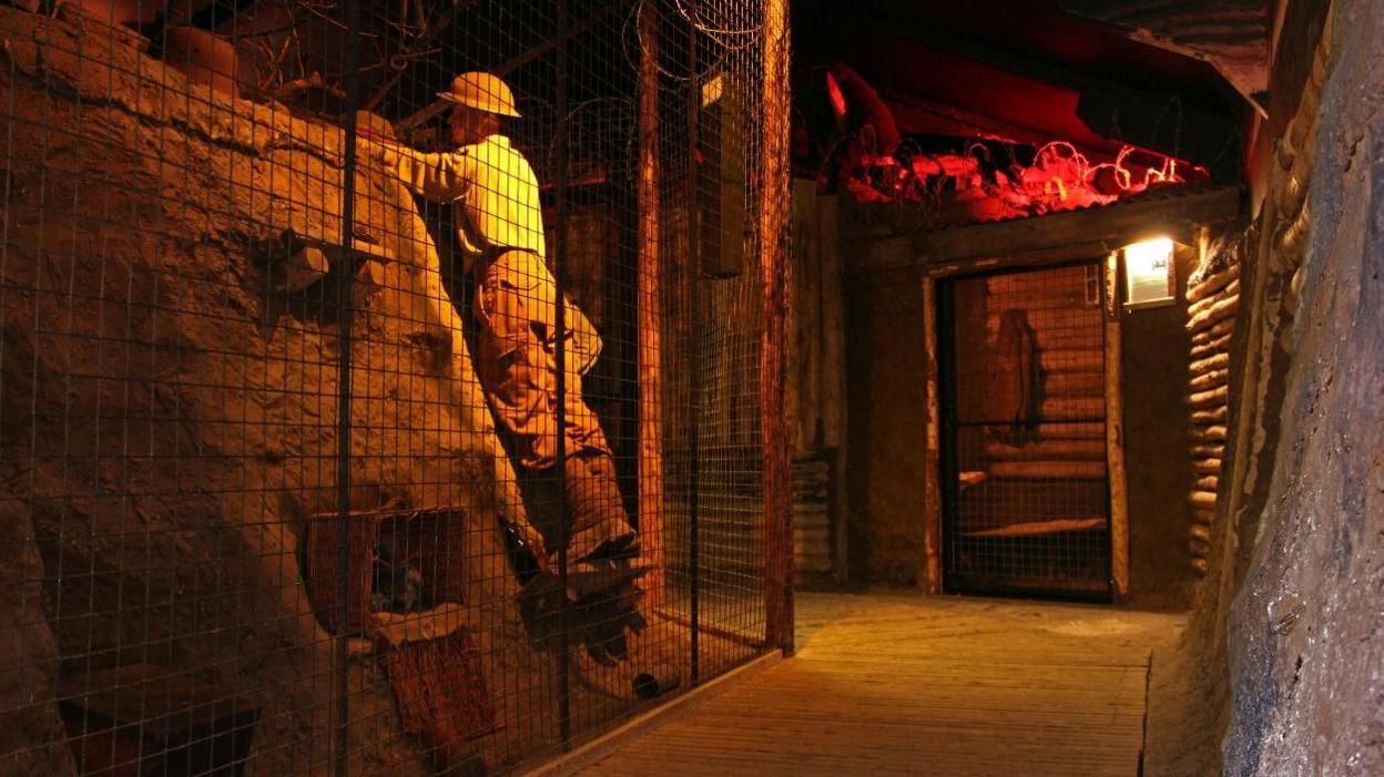 An exhibit showing a high fence on the left, a path in the middle and a grey wall on the right. Behind the fence is a life size model of a mud bank with a model of a man in a jumper and trousers climbing the mud mound. 