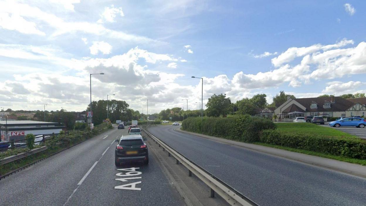 The image shows a two-lane road labelled A164 with traffic on one side, a metal guardrail dividing the lanes, a KFC on the left and a building with parking on the right under a sunny sky.