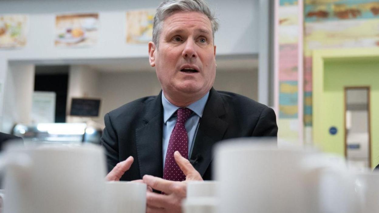 Sir Keir Starmer wearing a light blue shirt, maroon tie with dots and a dark blazer. His mouth is open, caught speaking mid-sentence, and he is gesturing with his hands