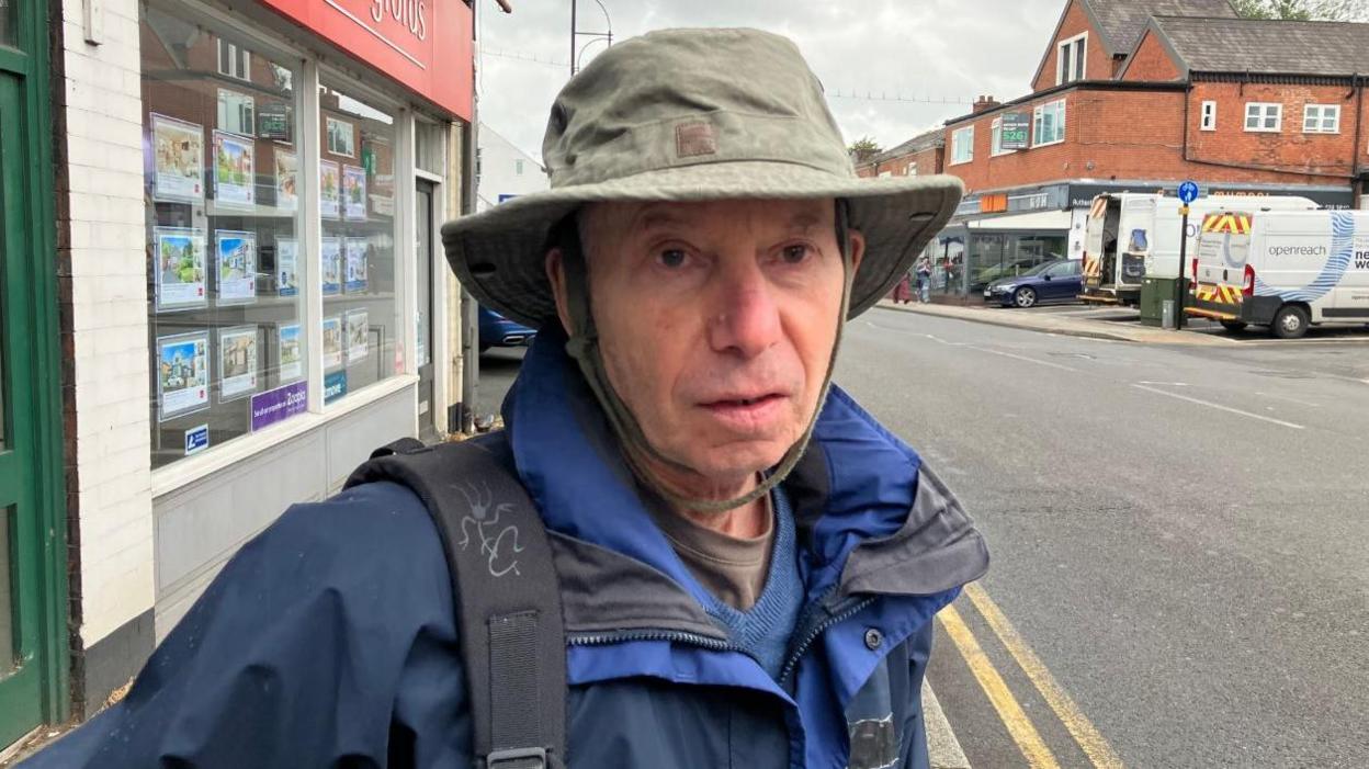 Dave Cooper is standing on the high street in Cheadle. Int the background is an estate agents. He is wearing a hat and waterproof coat along with a backpack.