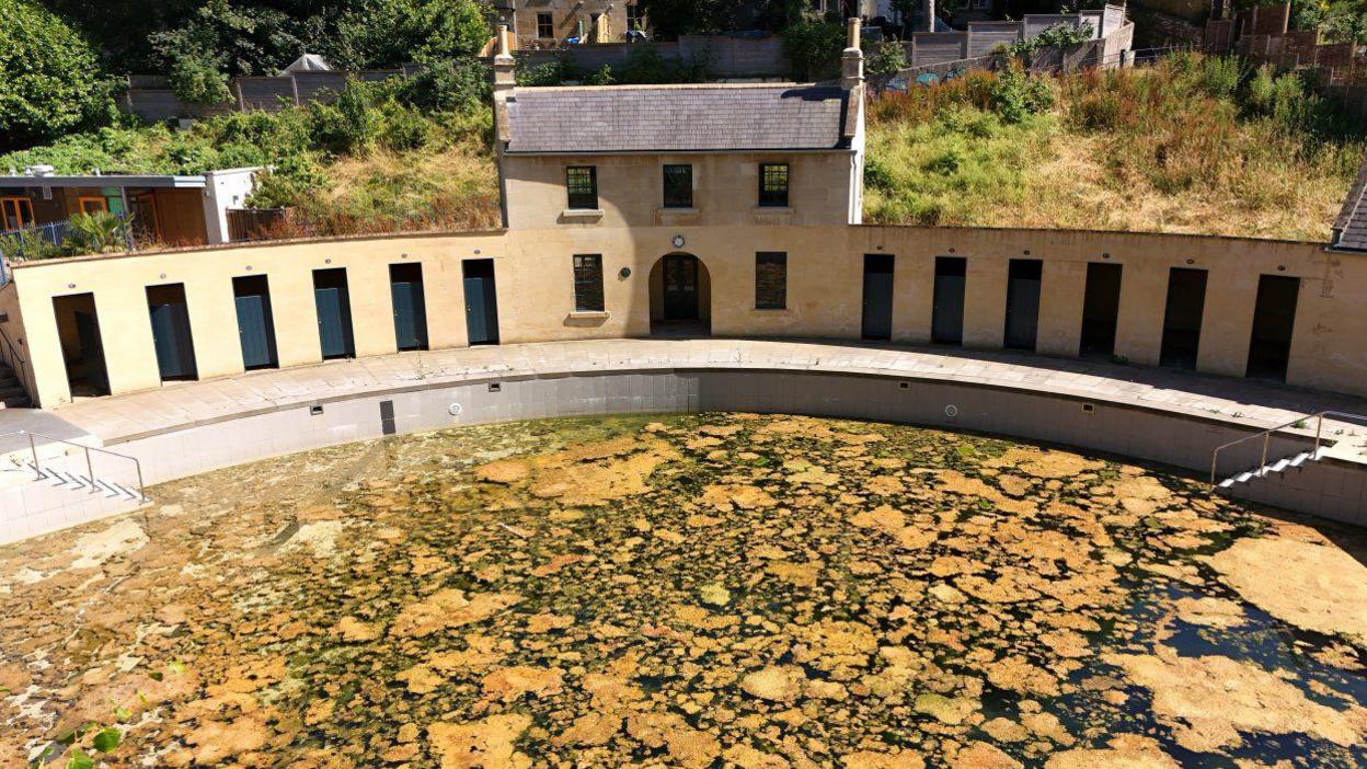 A drone image of Cleveland Pools, showing dark water with patches of what looks like yellow/brown algae. The Cotswold stone-style buildings are visible in the background