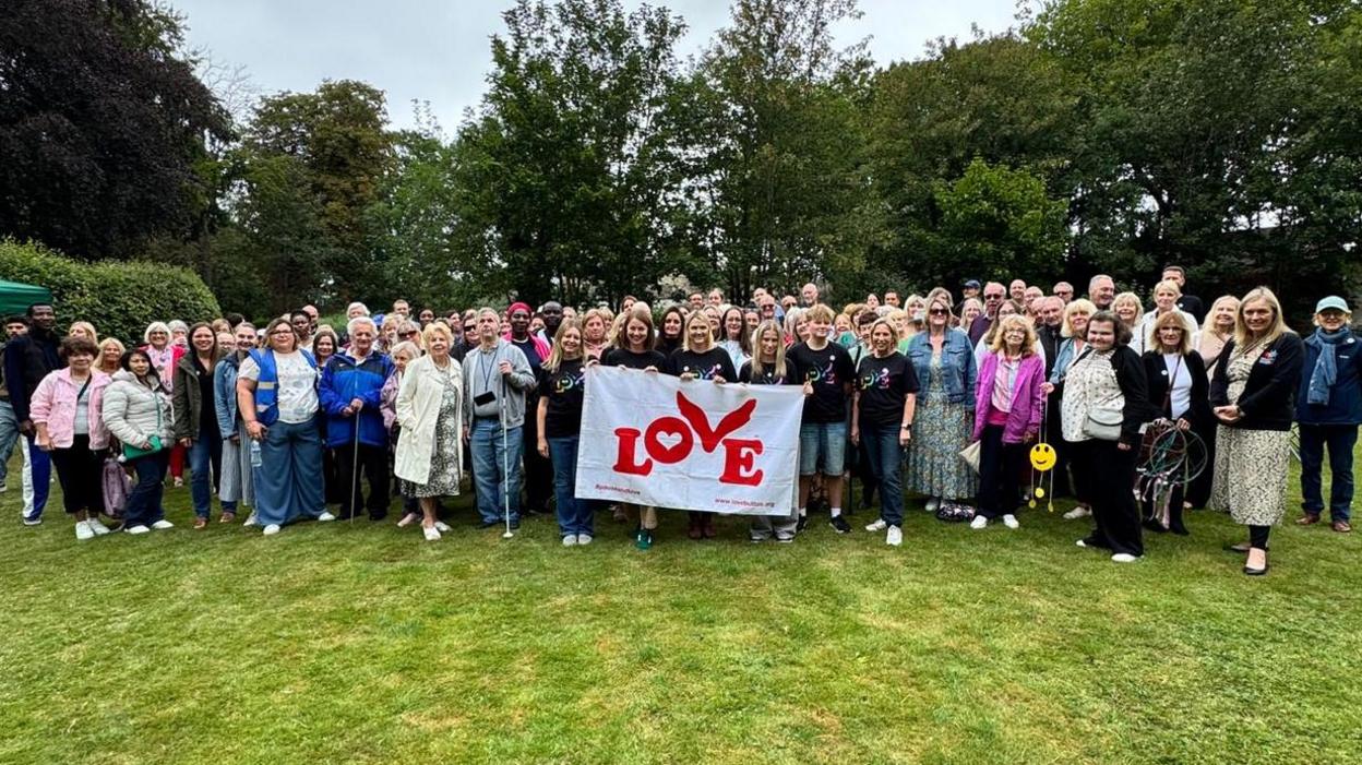 About 100 people holding a Coldplay love flag in the gardens at Holderness House. They're all smiling and grouped together.
