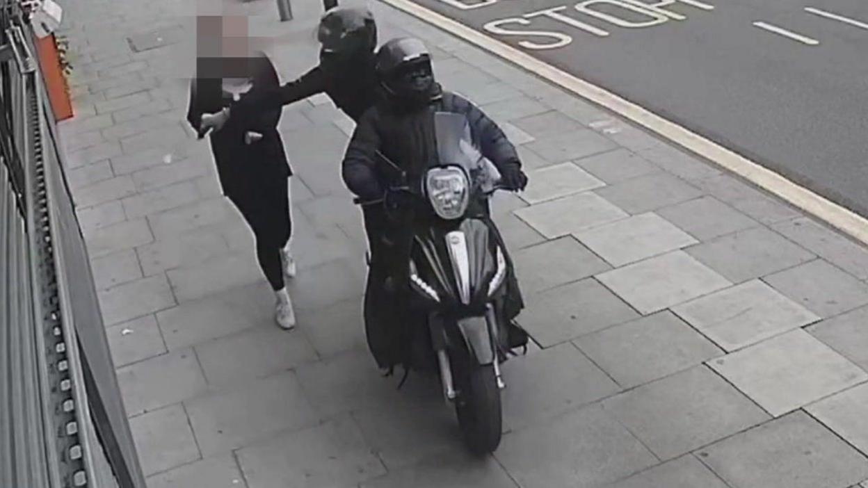 Two people on a moped are driving on the pavement past a pedestrian and one is reaching out to snatch the person's phone from their hand 