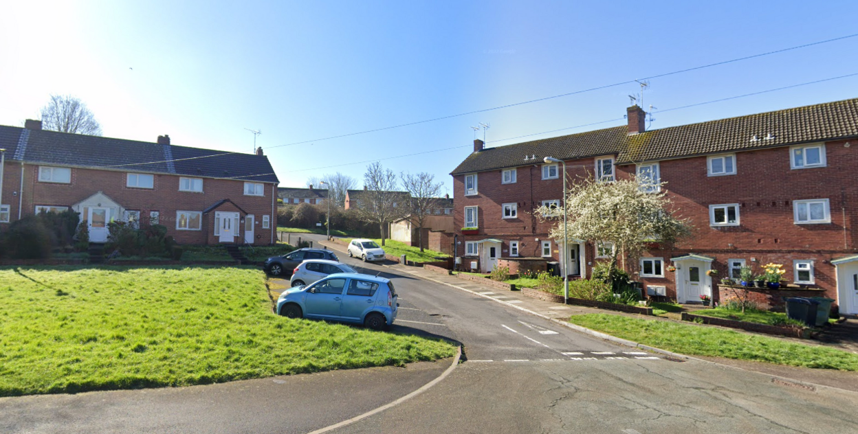 Google image of Durham Close in Exeter. Red brick terrace properties are on either side of the residential street. Cars are parked in parking bays outside of the properties. A green space is located outside of the homes. 