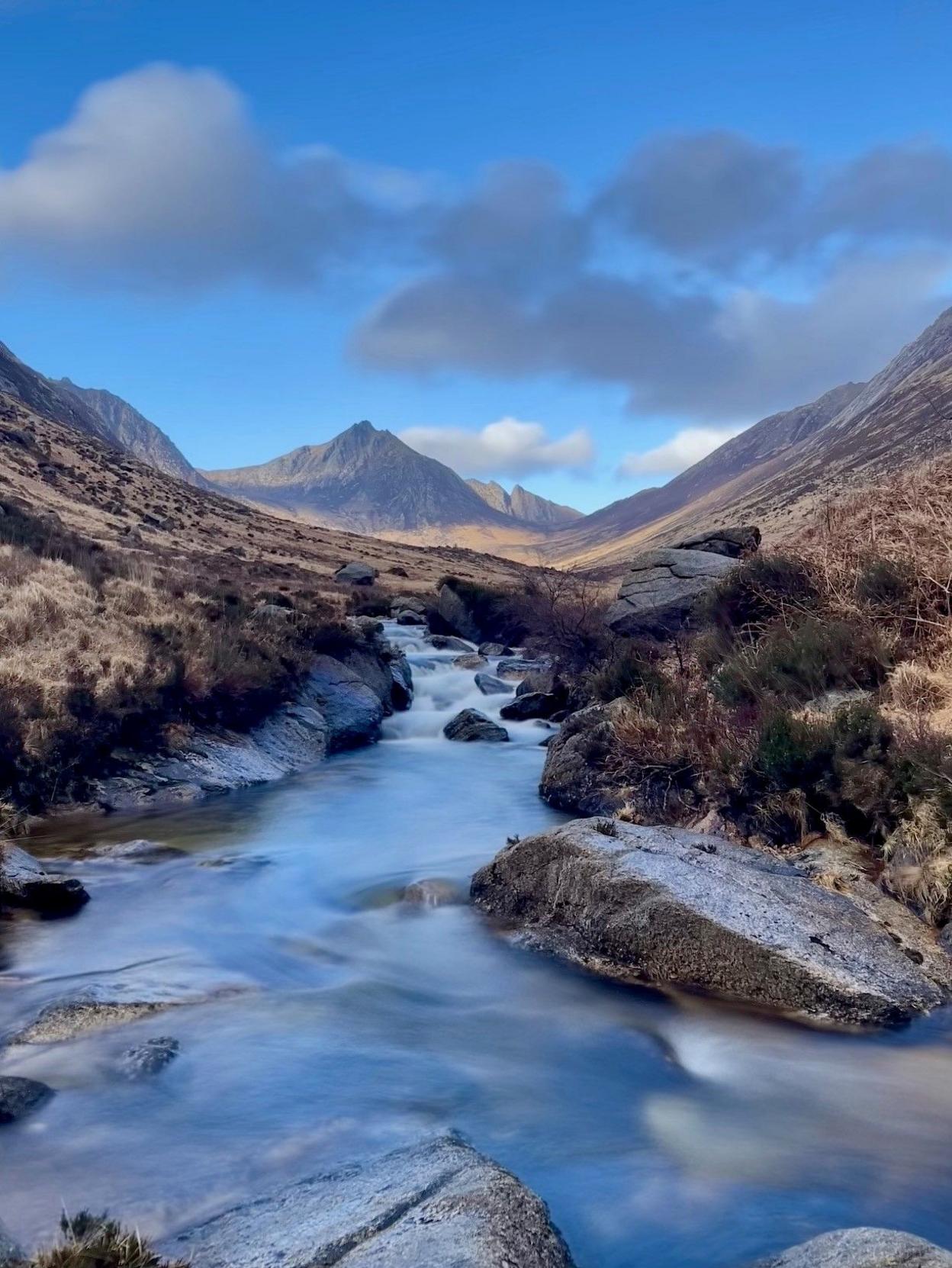 Portrait sized image of a small brook running through the valley between hills with a mountain in the distance and a few grey clouds against a bright blue sky