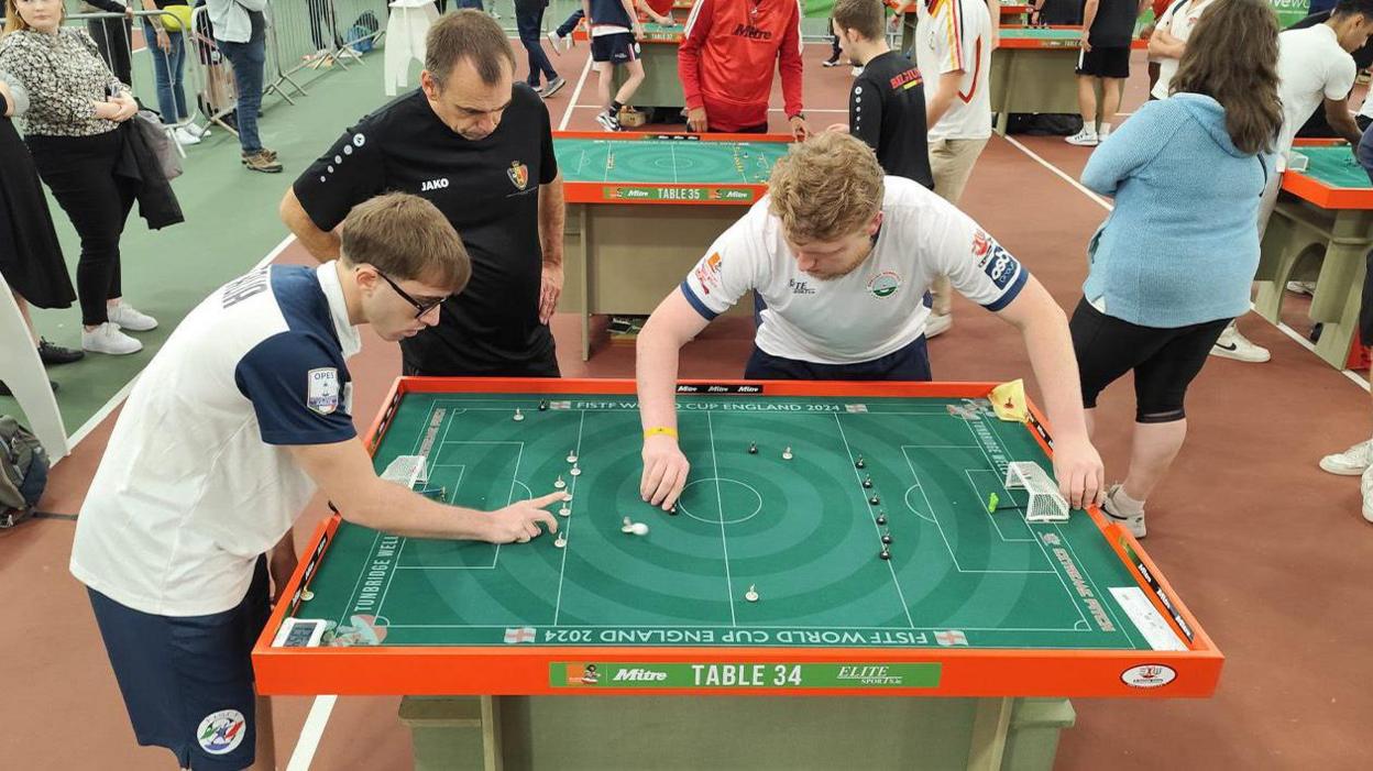 Two men wearing white shirts play Subbuteo as a referee wearing a black shirt watches on.