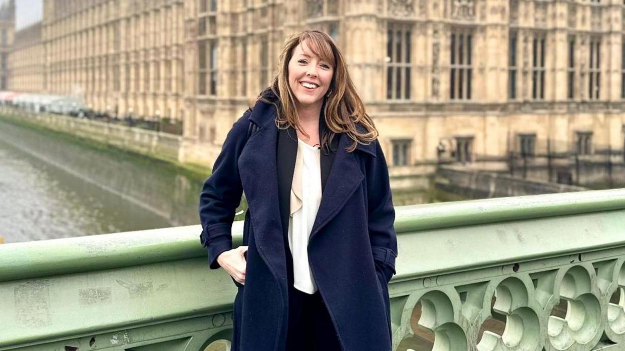 A woman with brown hair wearing a white shirt under a navy trench coat. She is standing in front of the Houses of Parliament.