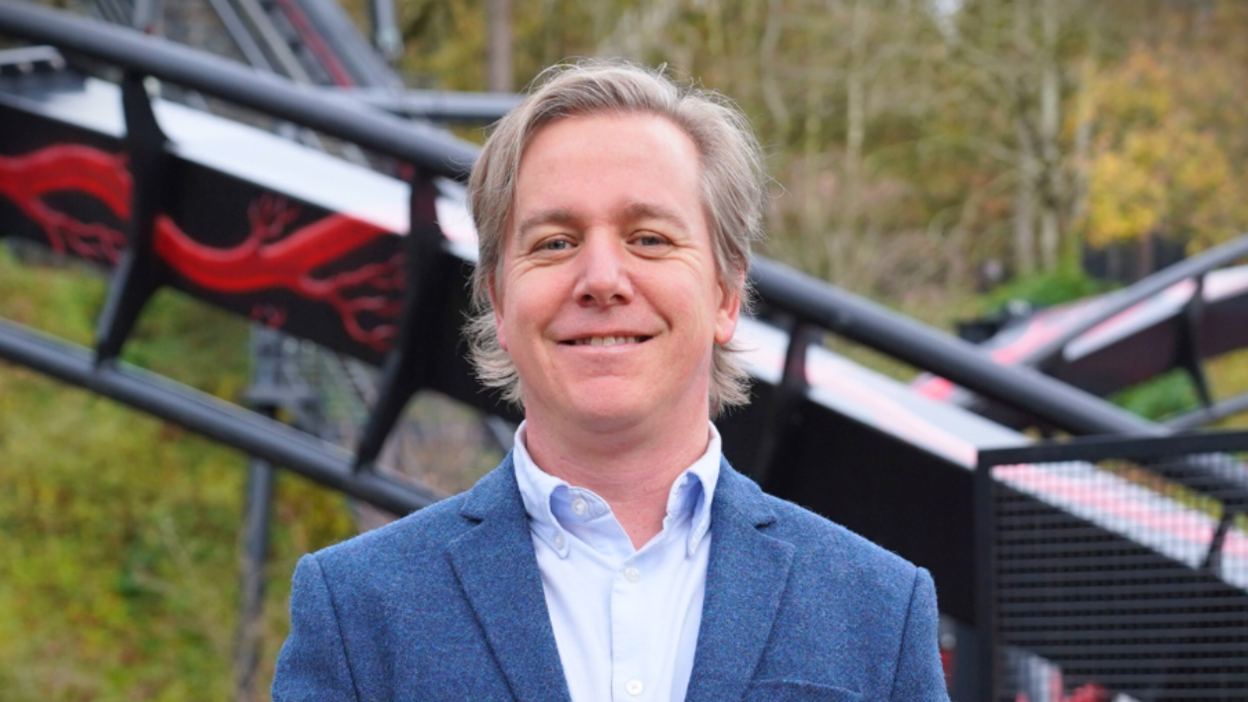 Neil Crittenden in a blue jacket and blue shirt standing in front of a section of the Nemesis Reborn rollercoaster at Alton Towers