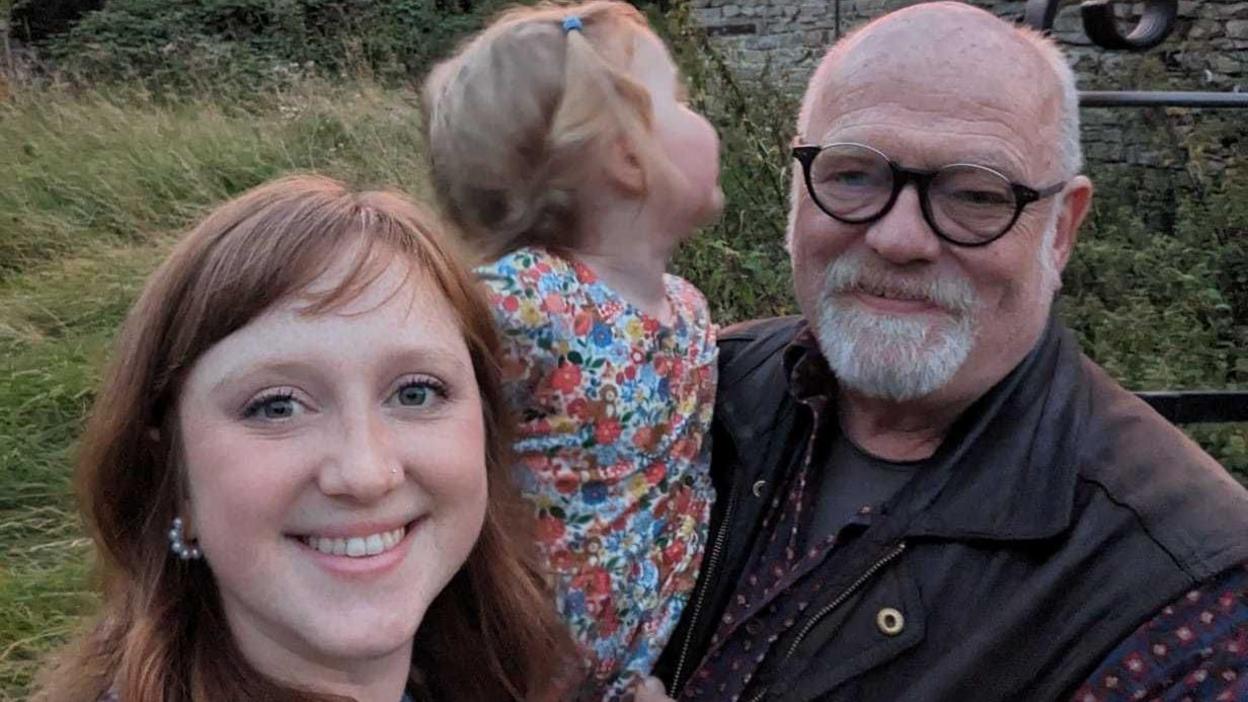 Cicely, pictured on the left, is smiling at the camera with a small child in the centre and her father on the right. She has red hair, cut into a fringe, and the child has blonde hair in pigtails and is wearing a multicoloured floral dress. Mr Berry is wearing a brown jacket or gilet with a t-shirt on. 