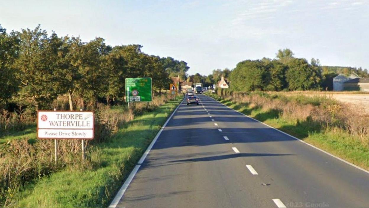 A road with cars and a lorry in the background and "Thorpe Waterville" sign