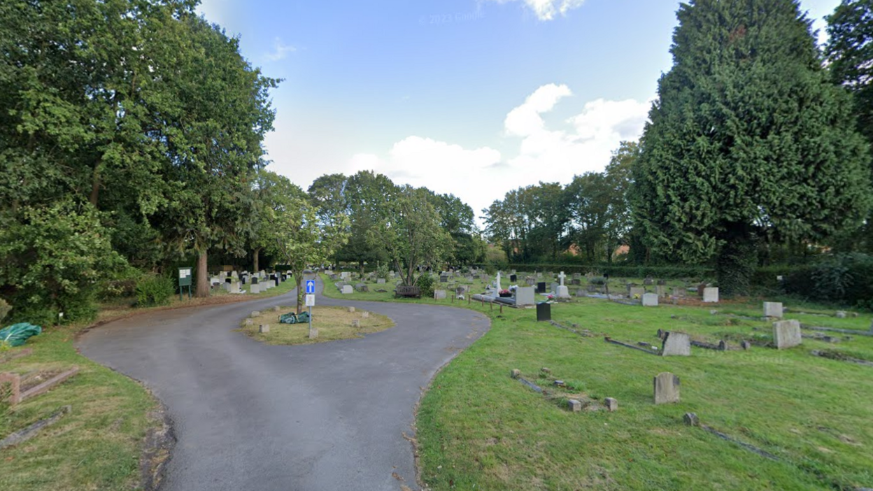 A screenshot from Google street view showing a cemetery with a road in the middle, with a small traffic island in the middle. There are trees on either edge of the cemetery and it's a cloudy day.