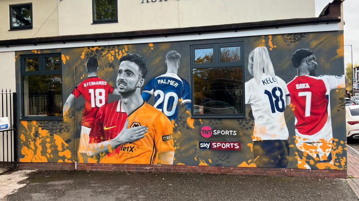 A mural of football players on a wall. It shows a football player with his hand on his chest, with his football shirt divided into two, representing two different clubs. Four other football players are behind him with their backs in view and not their faces.