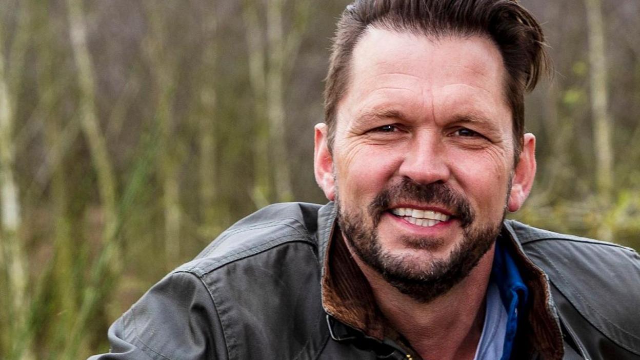 A photo of Jimmy Doherty looking at the camera. He is standing in a field with tall bushes pictured in the background behind him. He has dark hair which has been swept back atop his head. He is wearing a dark green coat with a jacket and top underneath.