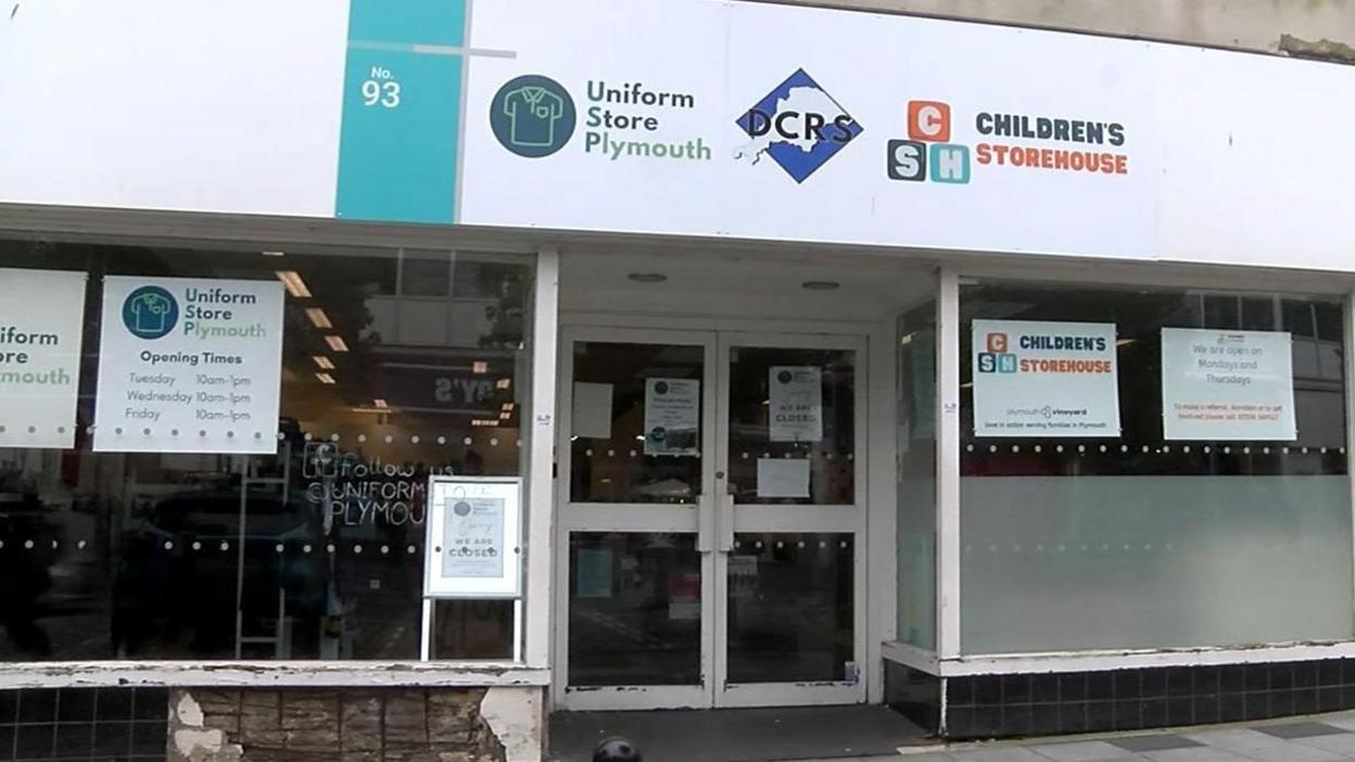 The outside of the building, which houses the hygiene hub. It is a slightly dilapidated building that appears to be a former retail shop. It has double doors and large windows either side. Tiles have fallen off the front of the building and the concrete is stained.  A large white sign stretching along the length of the shop shows the logos for two other groups offering children's school uniforms. Signs are in the windows showing the various opening times. 