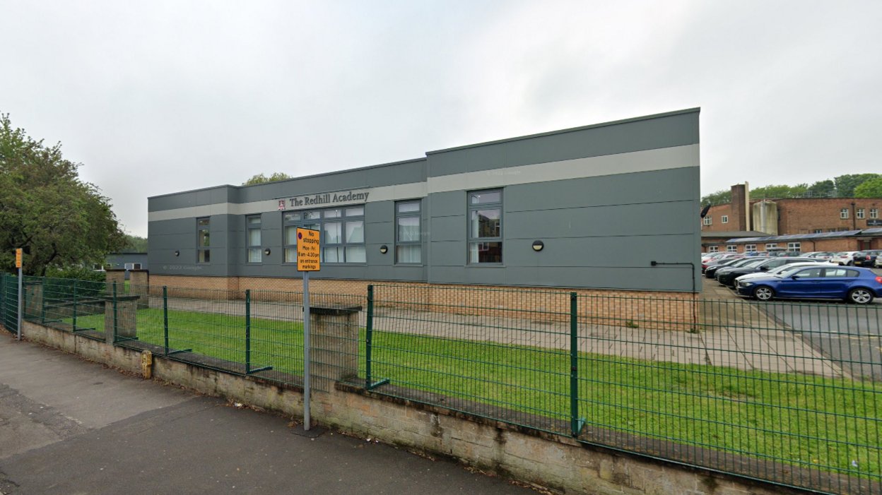 A grey single-storey school building behind a security fence 