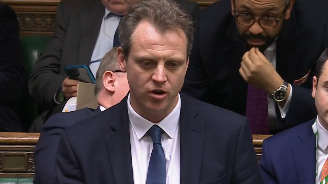 Joe Robertson is wearing a dark blue suit and blue tie and is talking to the House of Commons.
