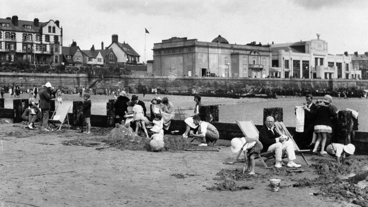 A black and white photo from 1955 of several people enjoying the beach at Bridlington. They are sitting on a breakwater windbreak or in deck chairs surrounding it. Children can be seen playing in the sand with adults sat around them. In the background you can see old buildings along the promenade.
