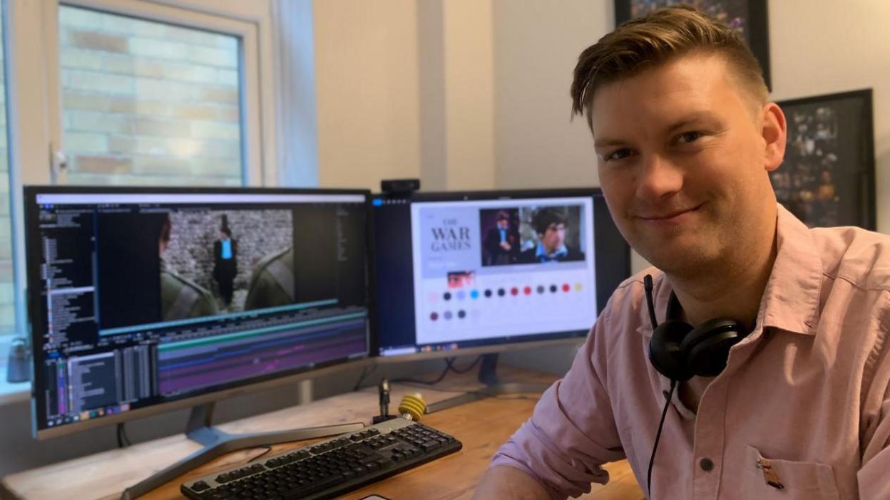 Rich Tipple smiling to camera wearing a pink shirt with two large monitors behind him showing a scene from the colour version of The War Games