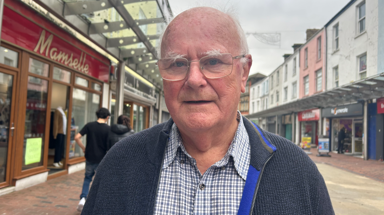 Edward Morgan, in a blue checked shirt and a navy cardigan. He has white hair and wears glasses, and is pictured standing in a street with shops and restaurants on either side.