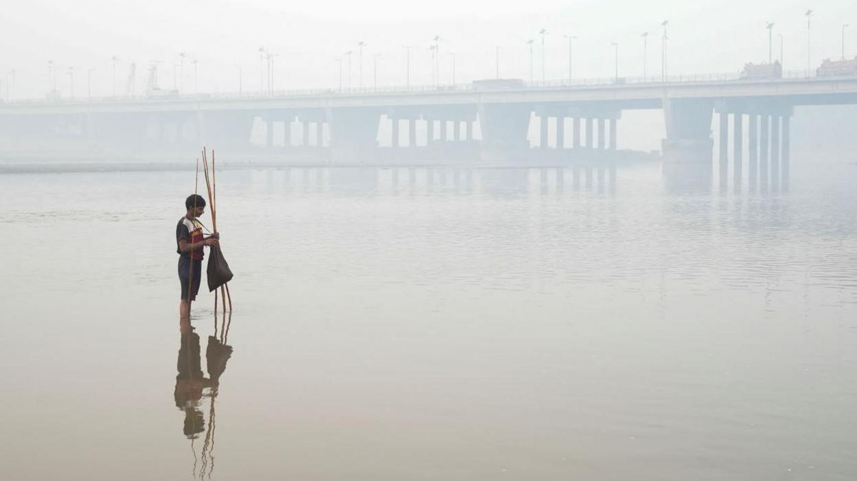 Young boy is setting up a fishing trap, with the bridge over the River Ravi in the background, amid smog in Lahore