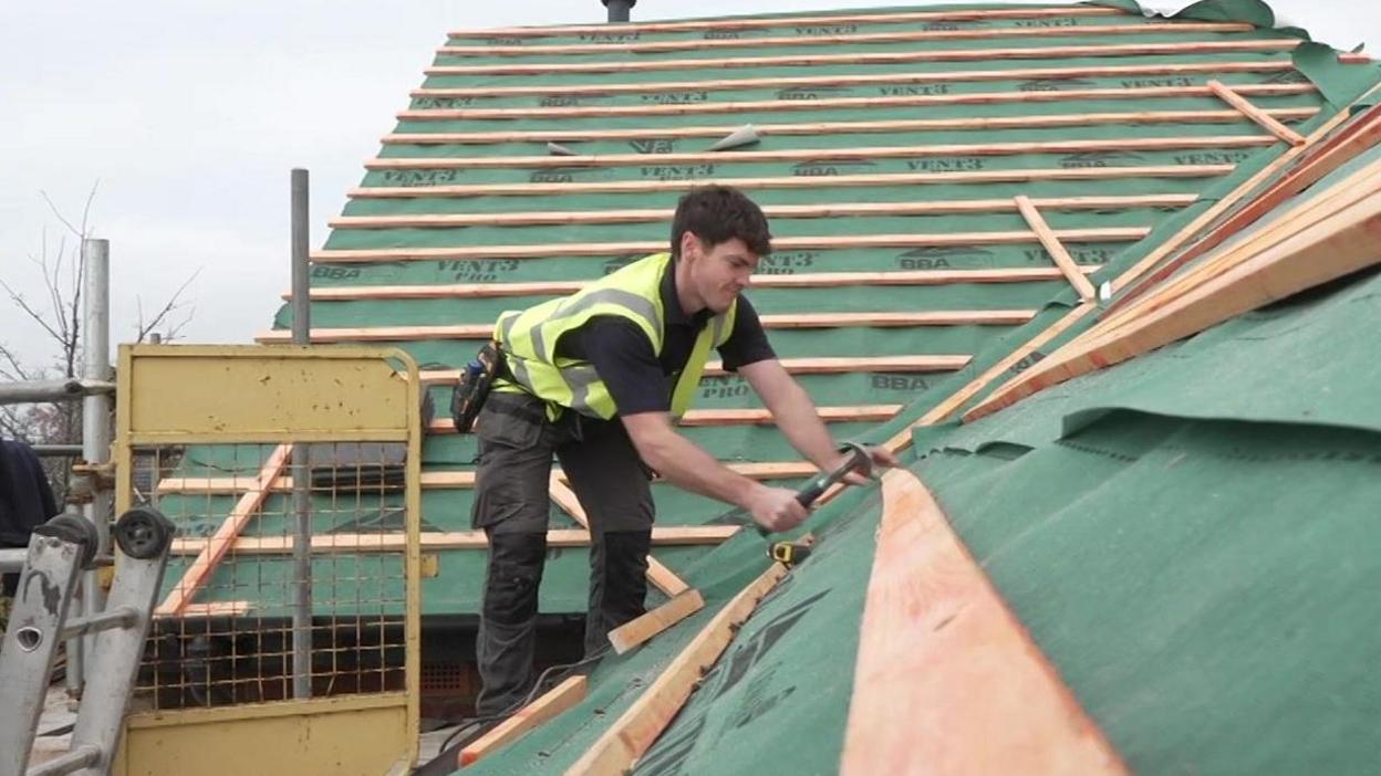 Joseph Gallagher hammering nails in to roof beams on top of a home on an overcast day. Joe is wearing a yellow high-vis vest. Surrounding him there are sheets of green roofing felt laid across two roofs, with wooden beams laid horizontally in rows.