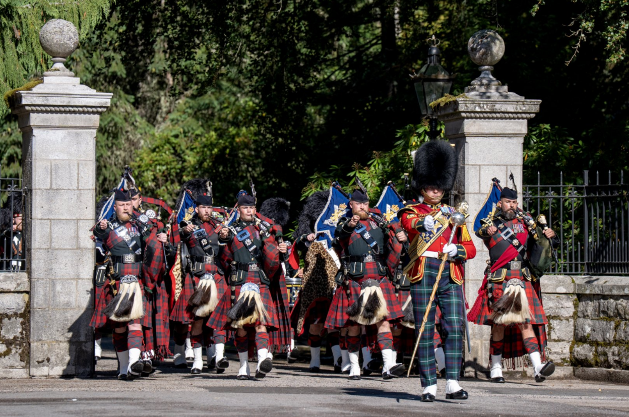 The Band of the 2nd Battalion marches through a stone gateway during a ceremonial parade. Musicians in tartan kilts and dark military uniforms play bagpipes while advancing in formation behind a drum major in a red tunic and bearskin-style hat. 
