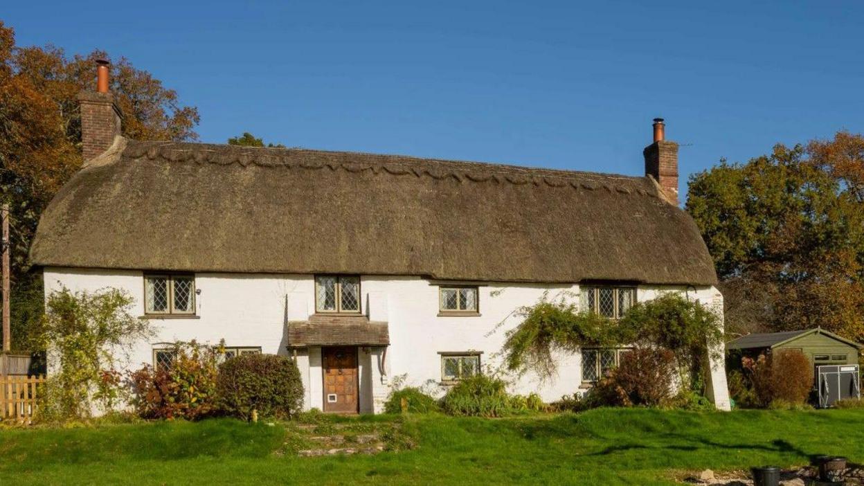 A pretty two-storey thatched cottage with white walls and eight lead-pane windows. It is surrounded by grass and trees.
