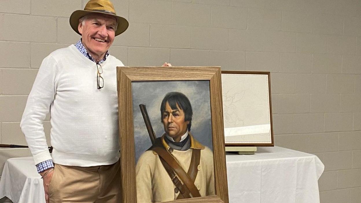 A middle aged man wearing a cream jumper and a hat, standing next to a portrait of David Thompson. D'Arcy Jenish is one of the leading authorities on David Thompson