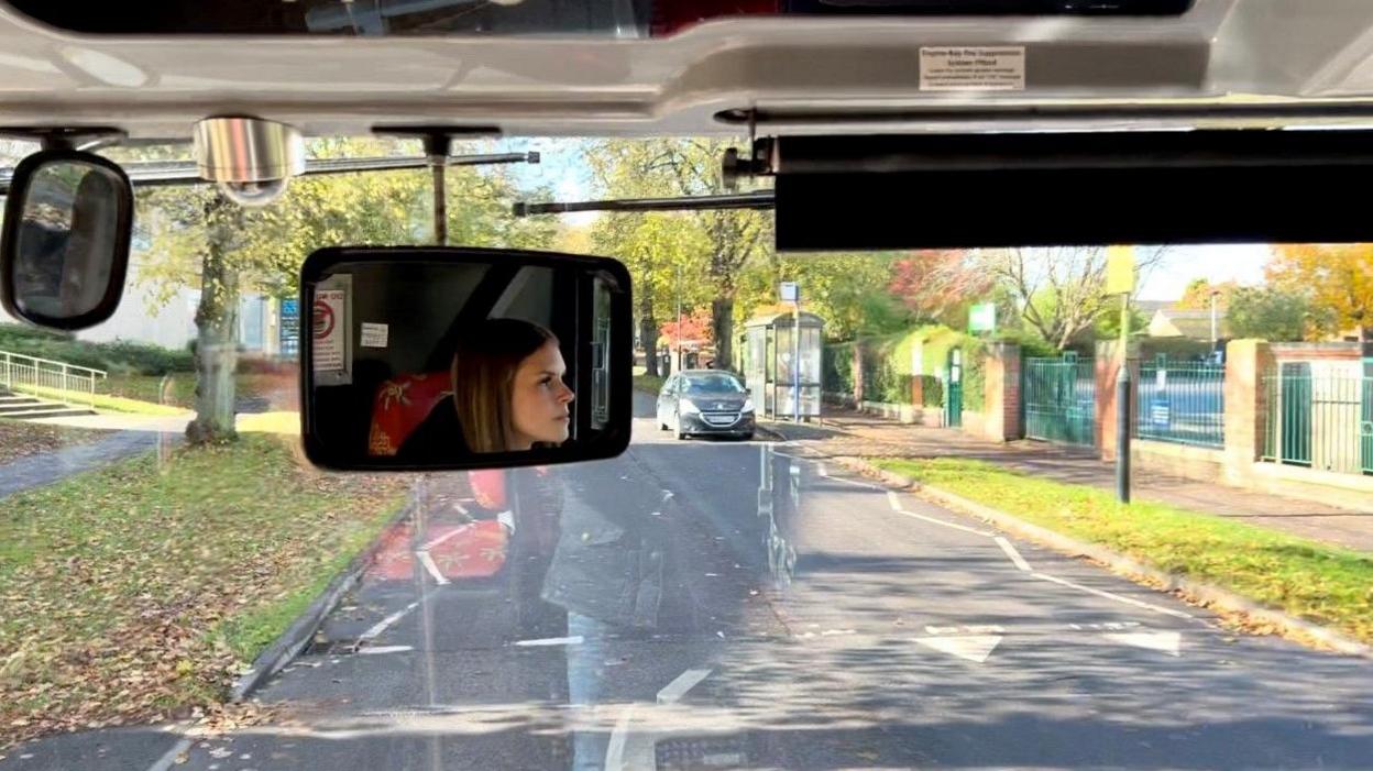 Emma Pool-Long's face is reflected in the rear view mirror while out of the windscreen is a road with grass verges on both sides.