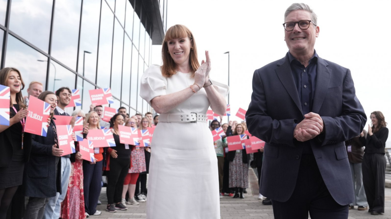 Sir Keir Starmer, wearing a dark suit, smiles next to Angela Rayner, dressed in white, as the two arrive in Liverpool ahead of Labour's Party conference. Against the backdrop of the two are Labour supporters who are smiling, applauding, cheering and holding up red Labor signs.