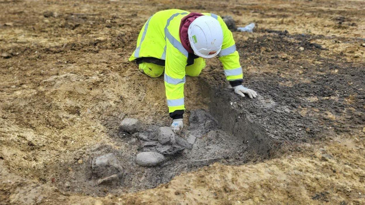 An person dressed in high-vis jacket and trousers is investigating charred items found on an archaeological dig.