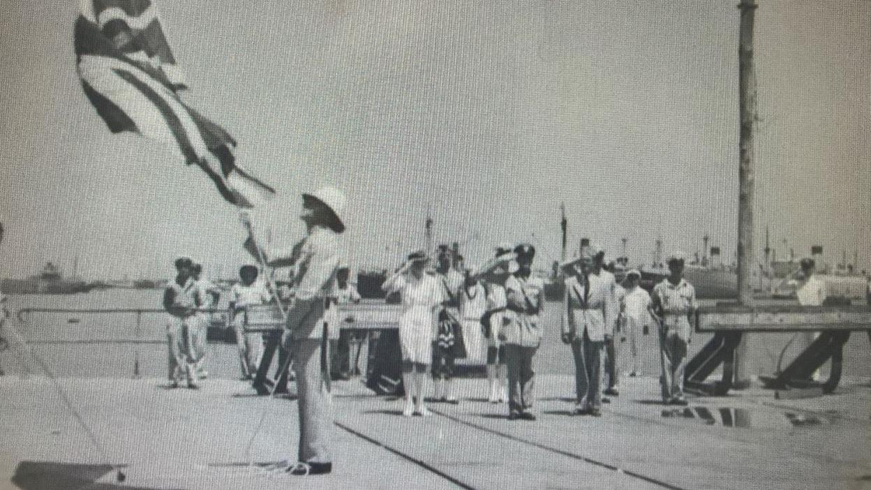 A black and white image of Tony Noakes in 1948. He is wearing a pith helmet and uniform as he lowers a British flag with warships in the background. Other military personnel stand behind him saluting. 
