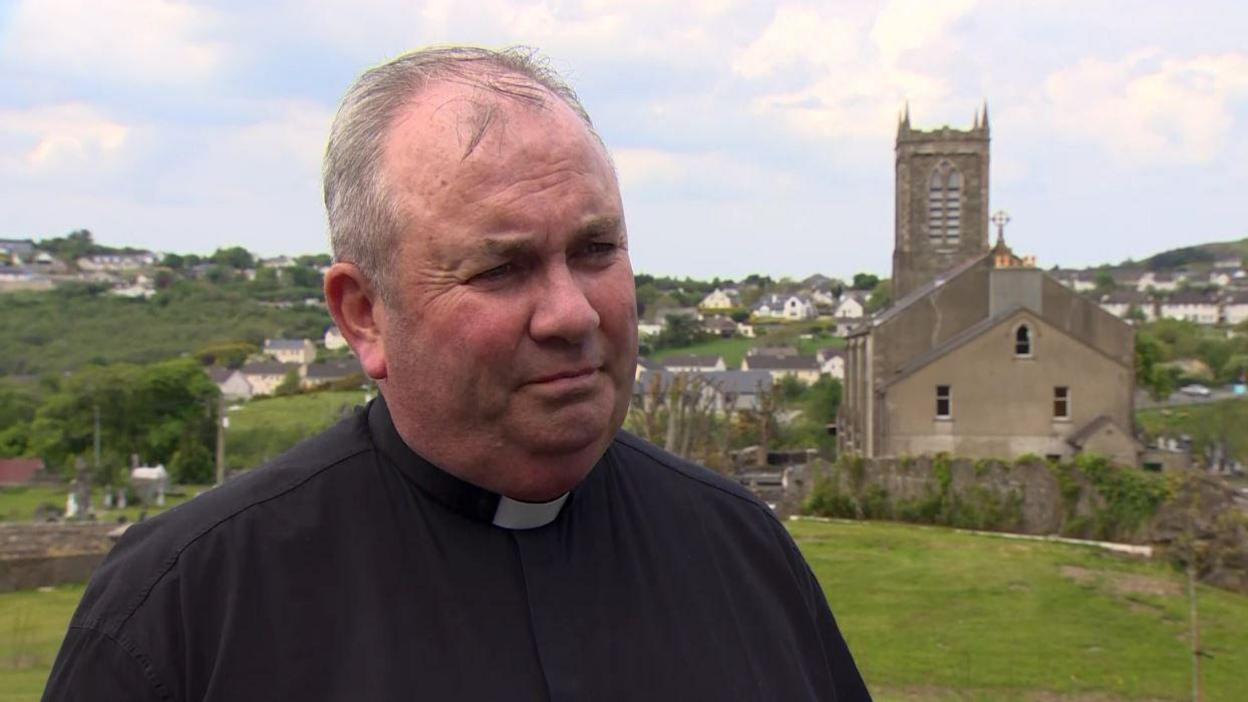 A priest, with short grey hair, looking at the camera. There is a field and a church in the background.