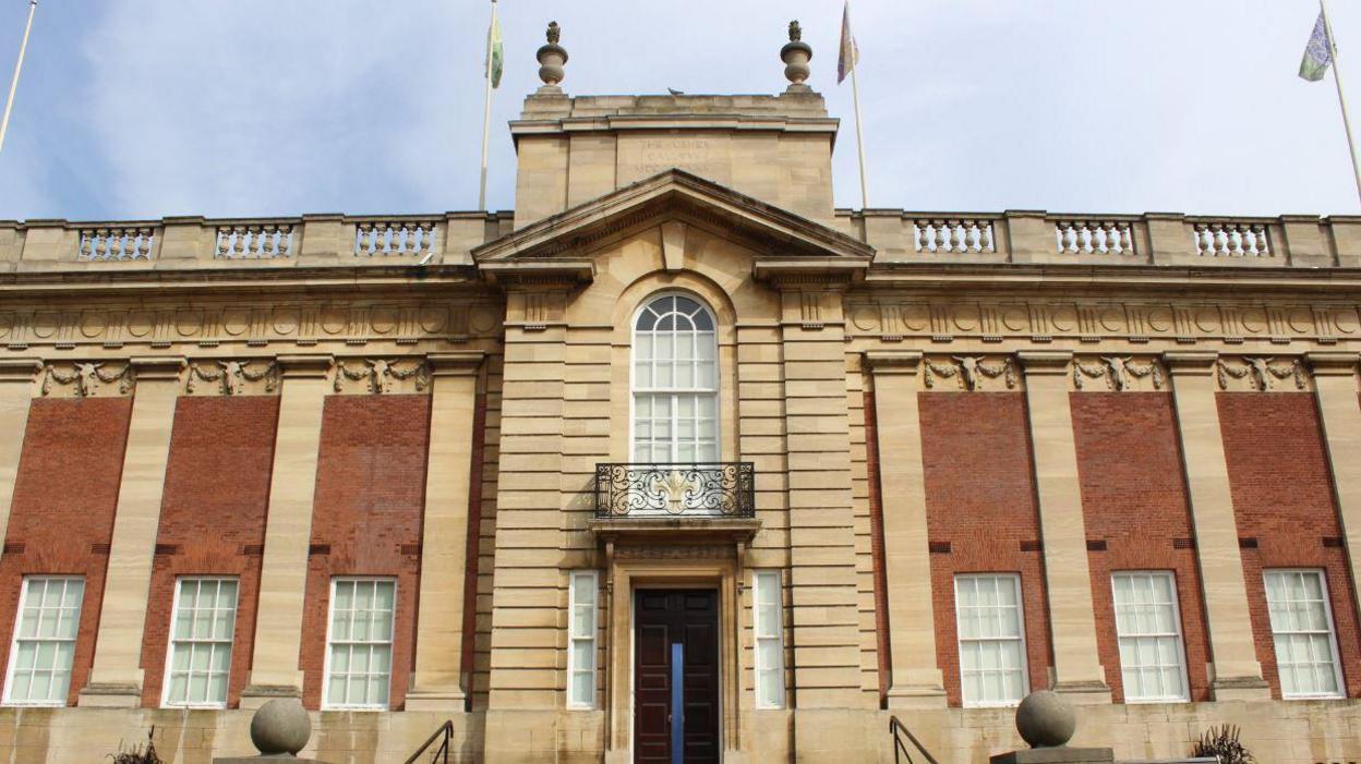 The exterior of the Usher Art gallery which has several stone pillars each side with windows in between. There is a black door in the middle for the entrance with a long white window and black balcony above it.