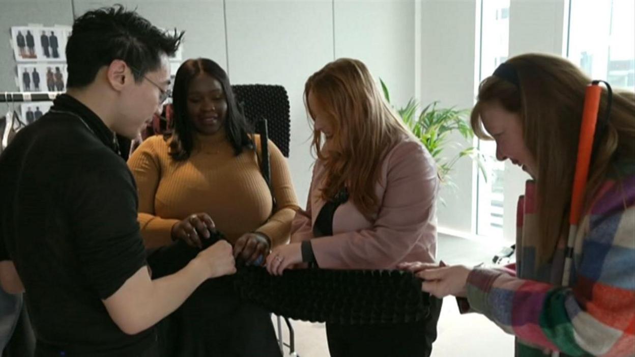 Fashion designer Chet Lo stands next to blind and visually impaired people Jessikah Inaba, Lucy Edwards and Jane Manley, who touch one of his black blazers made from textured pleated fabric 