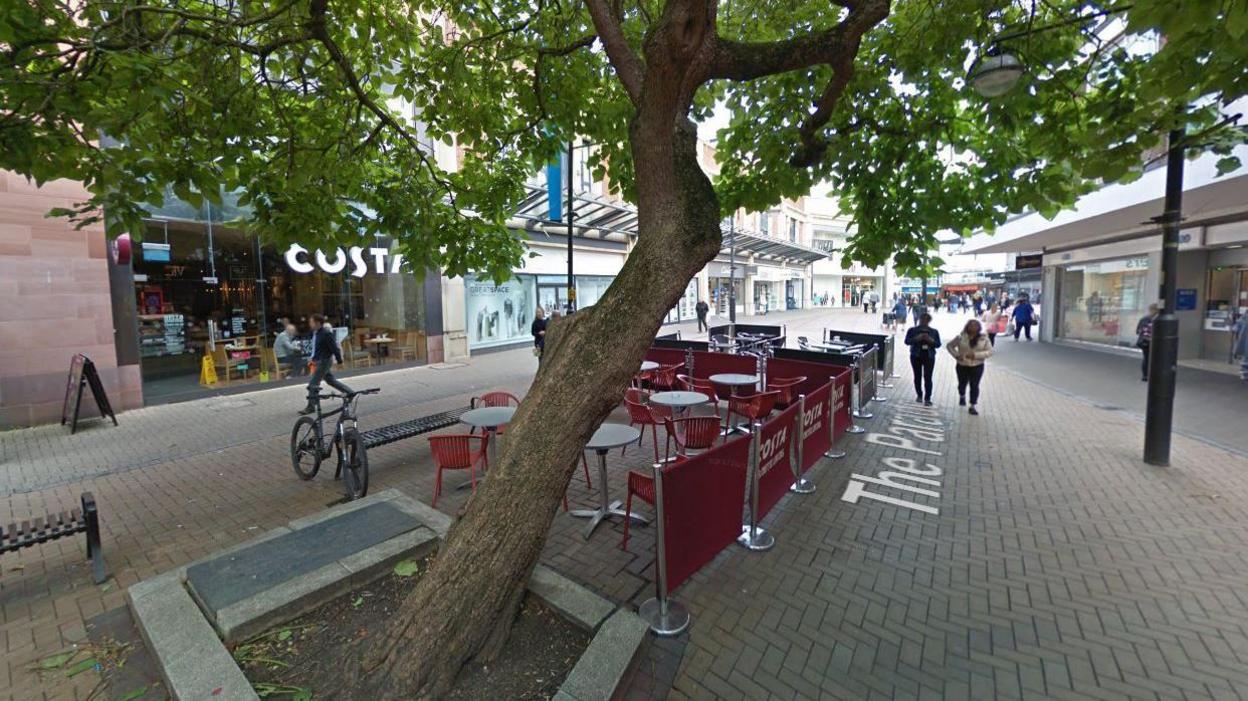 A google maps screenshot of the tree in the Parade. It is a shopping centre lane with a Costa coffee visible in the background and an outdoor seating area set up below the tree. The tree itself is leaning to the right at a 45 degree angle and has lots of leaves.