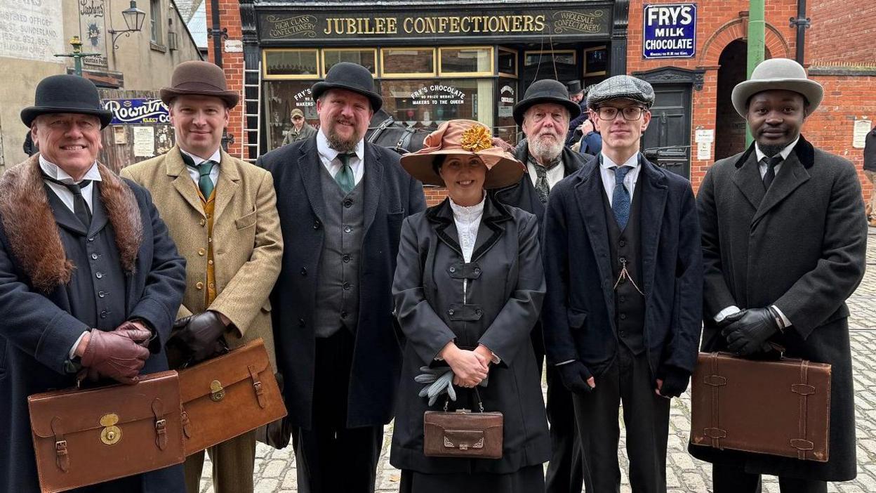 A group of male and female extras on the set of Night and Day at Beamish. They are in costume with the men wearing bowler hats or caps and suits and the woman wearing a wide hat a jacket and a dress. Most are carrying briefcases