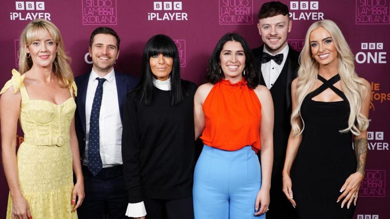 The finalists and host of The Traitors standing smiling at the camera during a photocall at BBC's New Broadcasting House. Left to right: Francesca Rowan-Plowden wearing a yellow dress, Alexander Dragonetti wearing a dark blue suit with spotted tie, Claudia Winkleman wearing a long black outfit with white ruffled collar and cuffs, Charlotte Berman in an orange/red vest top and sky blue trousers, Jake Brown wearing a black dinner jacket, waistcoat and bow tie, and Leanne Quigley wearing a long black dress.