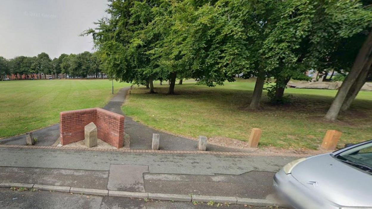 The edge of Peace Park, with a silver car parked on a street, close to bollards at the end of a pathway