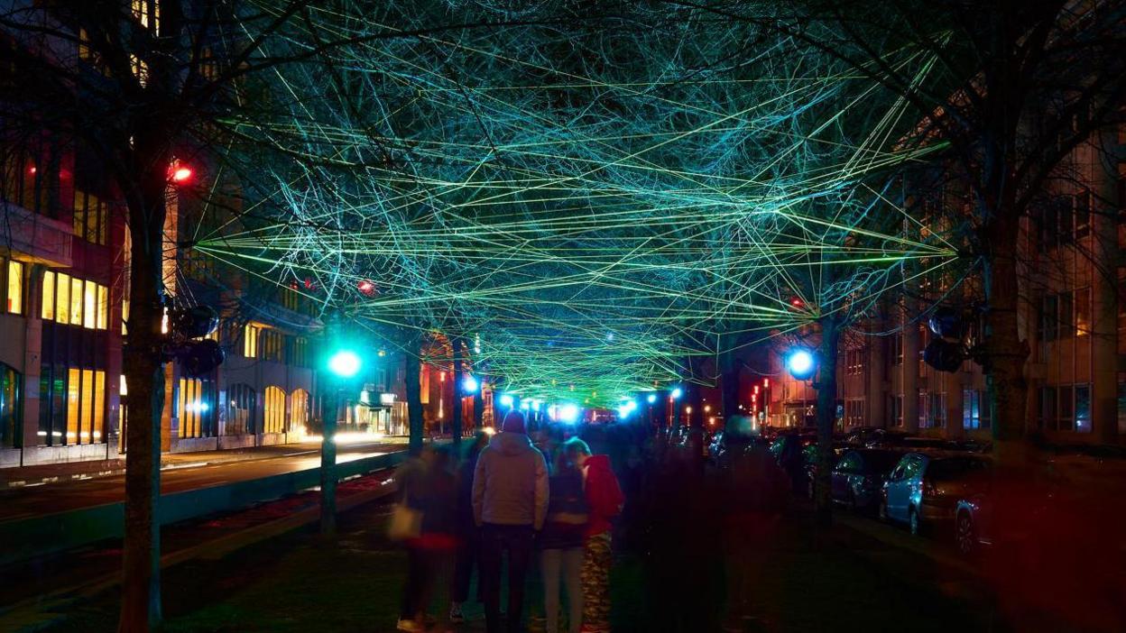 A group of people enjoy an intricate light installation is strung between trees on a city street.