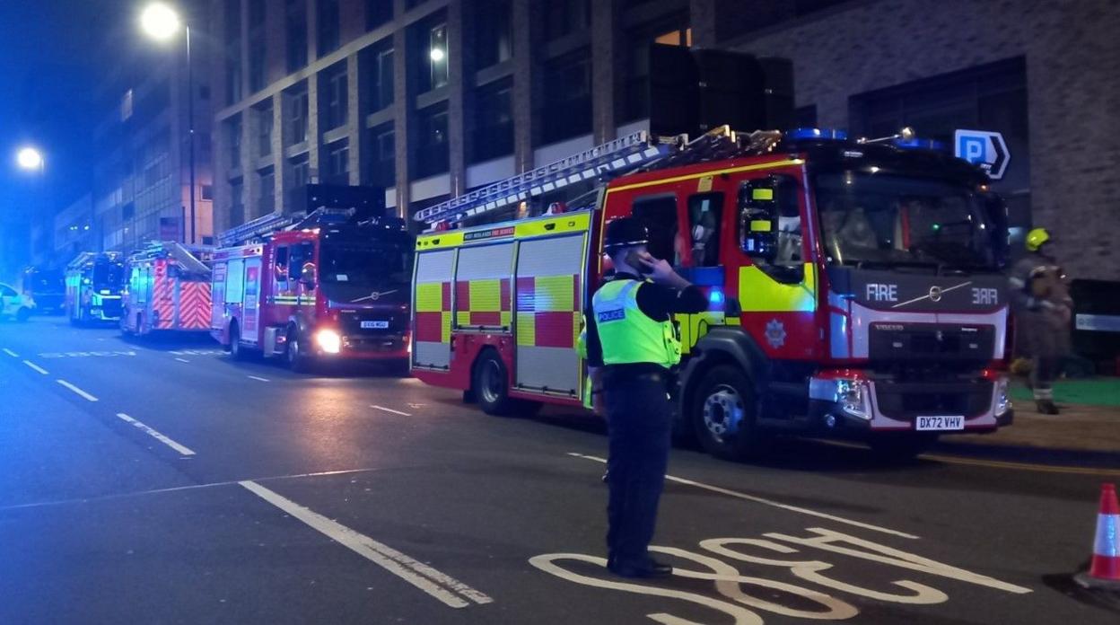 Five fire engines on one side of the road in the dark. A police officer, a cone and another vehicle are also on the street.