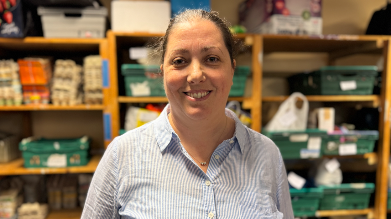 Erica Irwin has dark hair tied back and she's wearing a light blue shirt. She is standing in front of rows of shelves filled with green crates stocked with food boxes and bags.