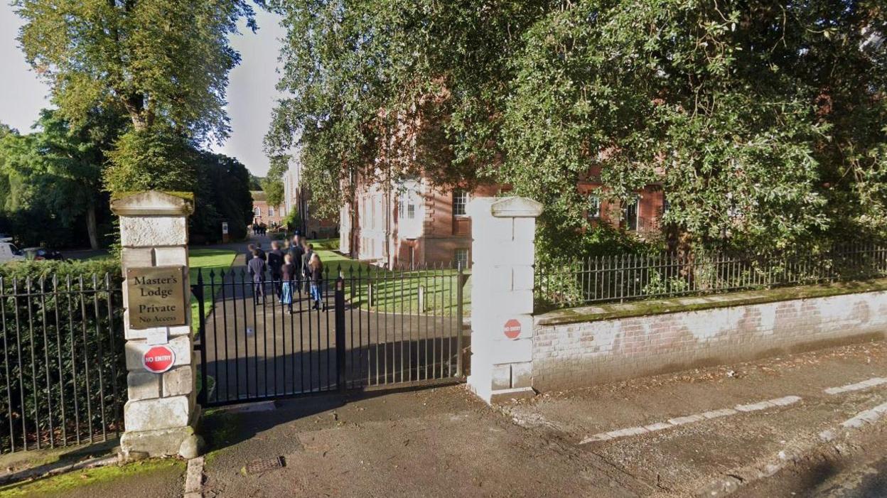 School gates in the foreground with the words: "Master's Lodge, Private, No Access" written on a plaque. School buildings and trees in background with pupils walking along the path away from the camera